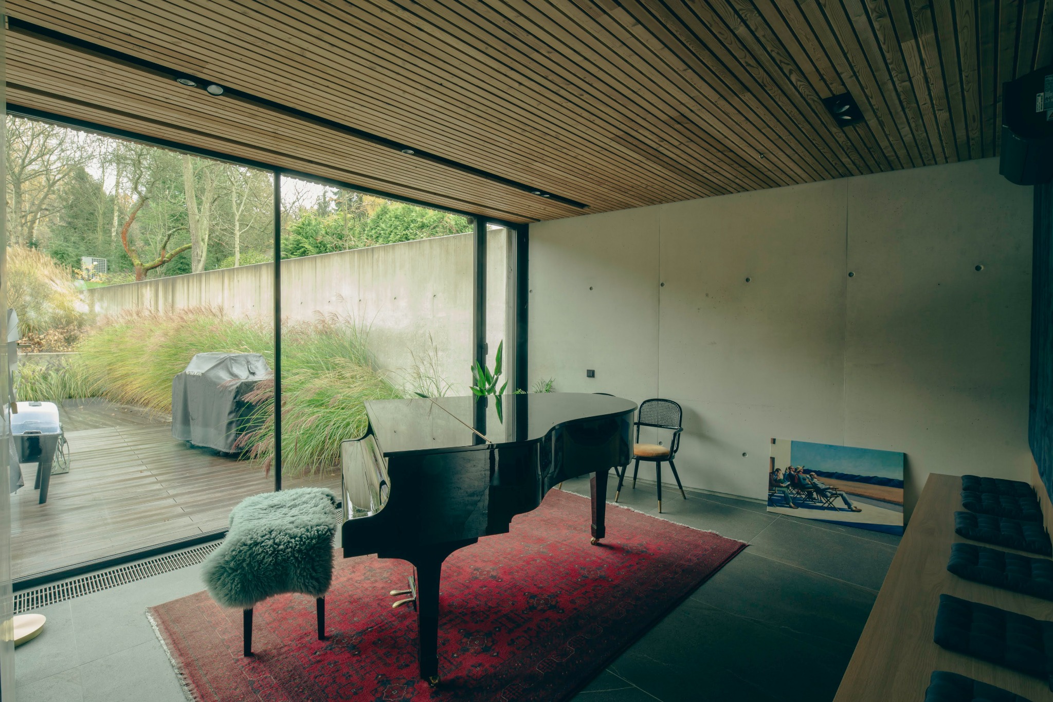 Nowoczesne wnętrze z czarnym fortepianem na czerwonym dywanie, widok na taras z trawami ozdobnymi i grillem, drewniany sufit lamelowy.