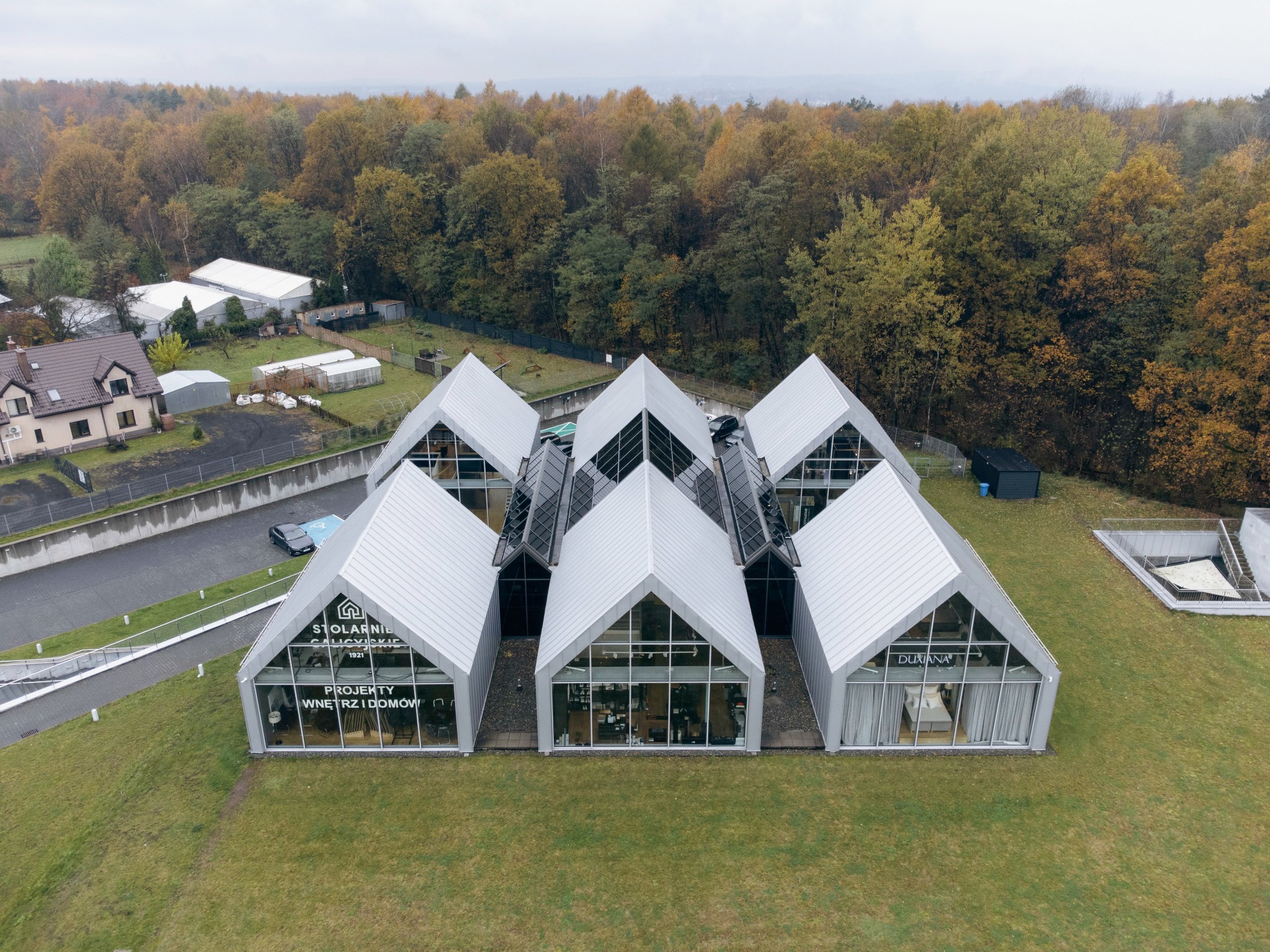 Widok z lotu ptaka na nowoczesny budynek z wieloma szczytowymi segmentami, przeszklonymi fasadami i metalowym dachem, otoczony zielenią i lasem w tle.