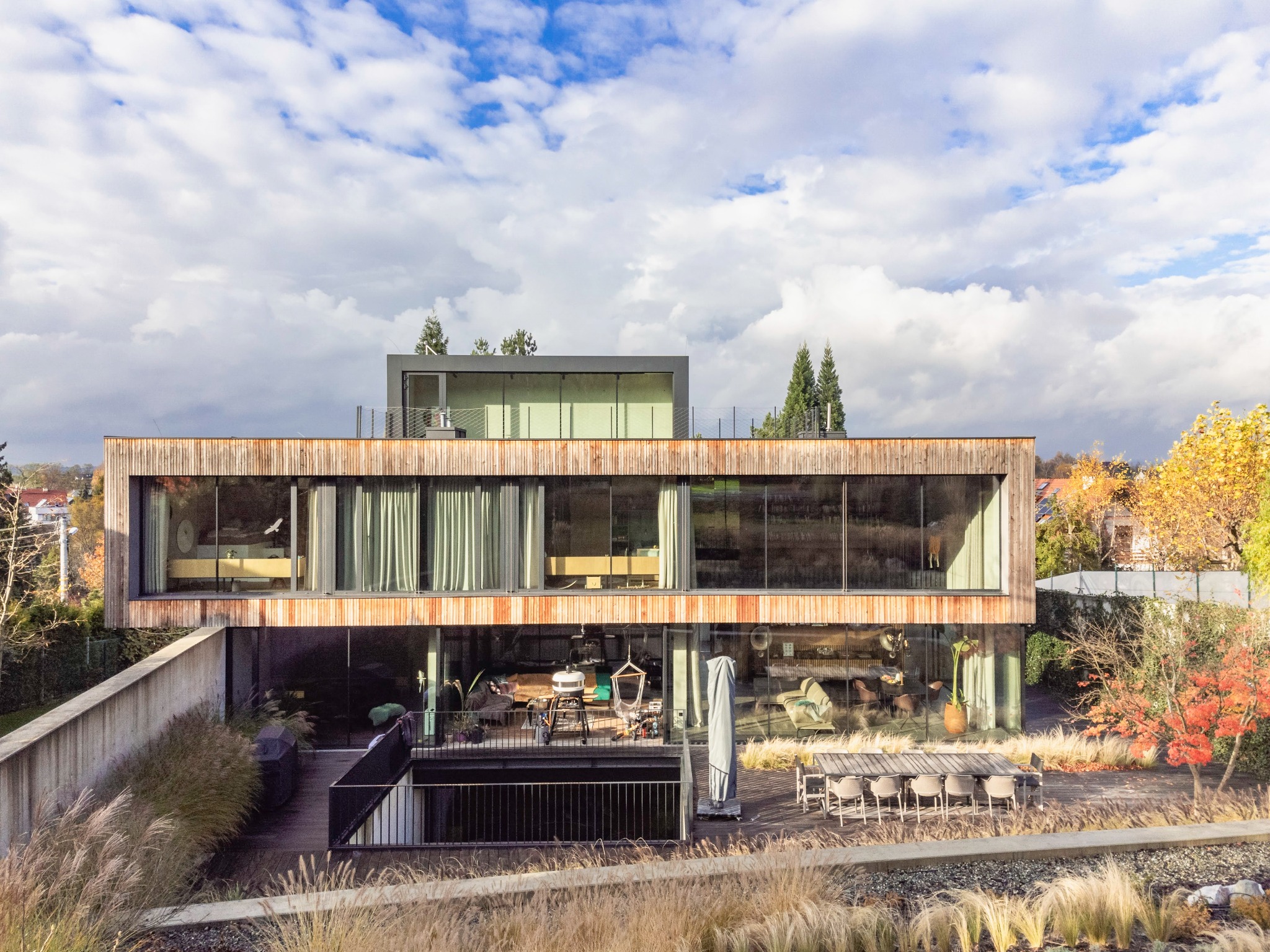 Nowoczesny dom z drewnianą elewacją i dużymi oknami, taras z meblami ogrodowymi i ozdobną trawą na pierwszym planie, widok z zewnątrz w słoneczny dzień.