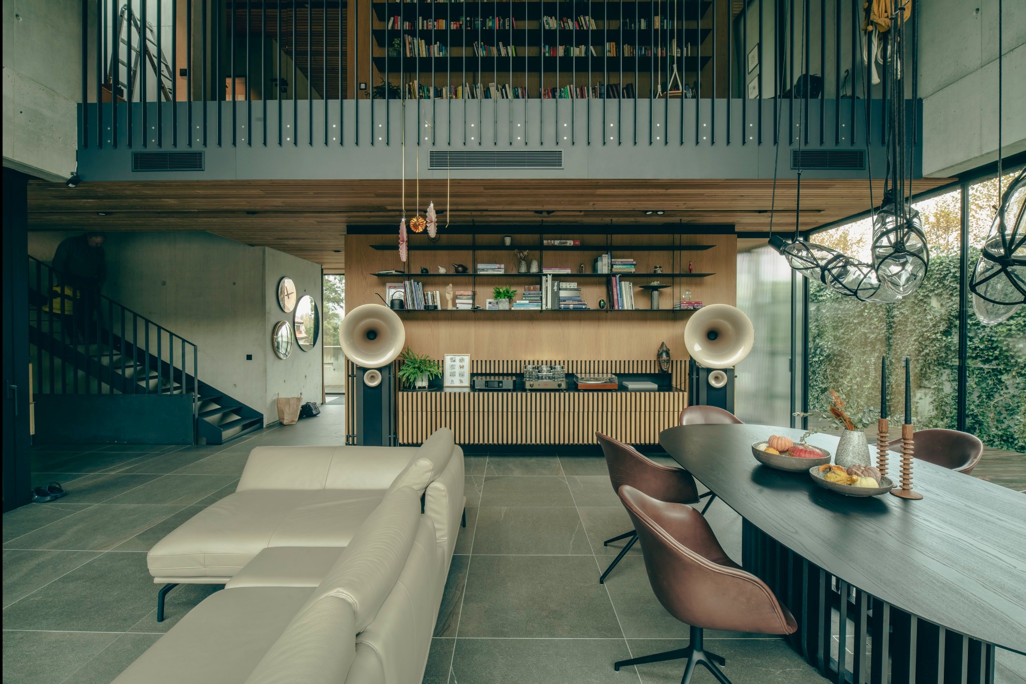 Przestronne wnętrze domu z antresolą, betonowymi ścianami, jasną sofą, drewnianą zabudową z półkami i gramofonem, oraz stołem z owocami w jadalni.