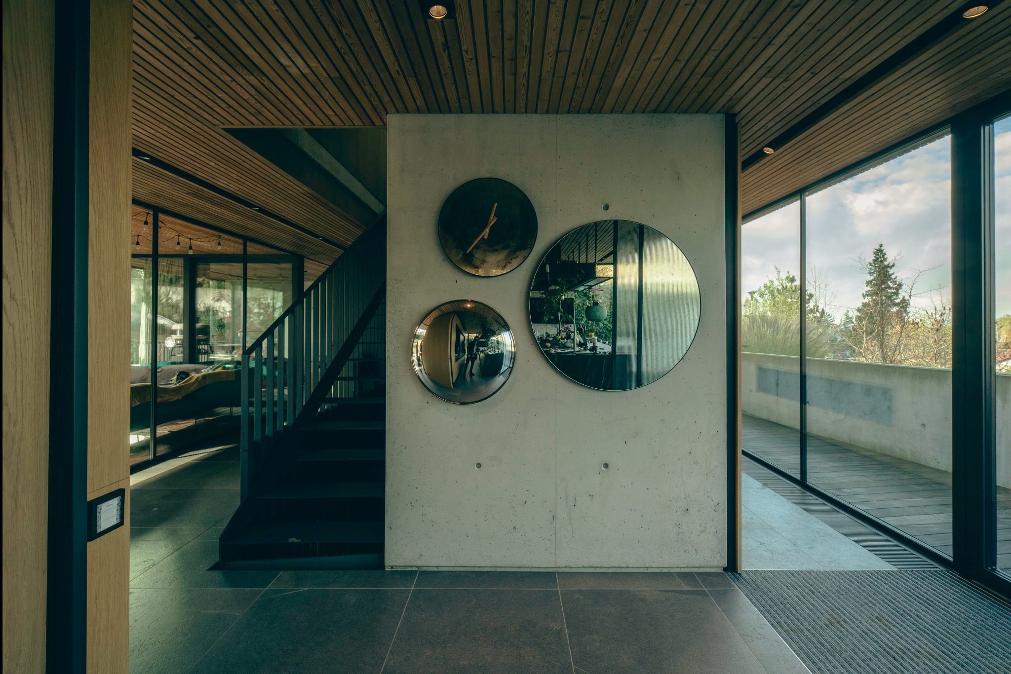 Nowoczesny hol z betonową ścianą, na której wiszą trzy okrągłe lustra i zegar, drewniany sufit z listew, schody z metalową balustradą i widok na zewnątrz przez duże okno.