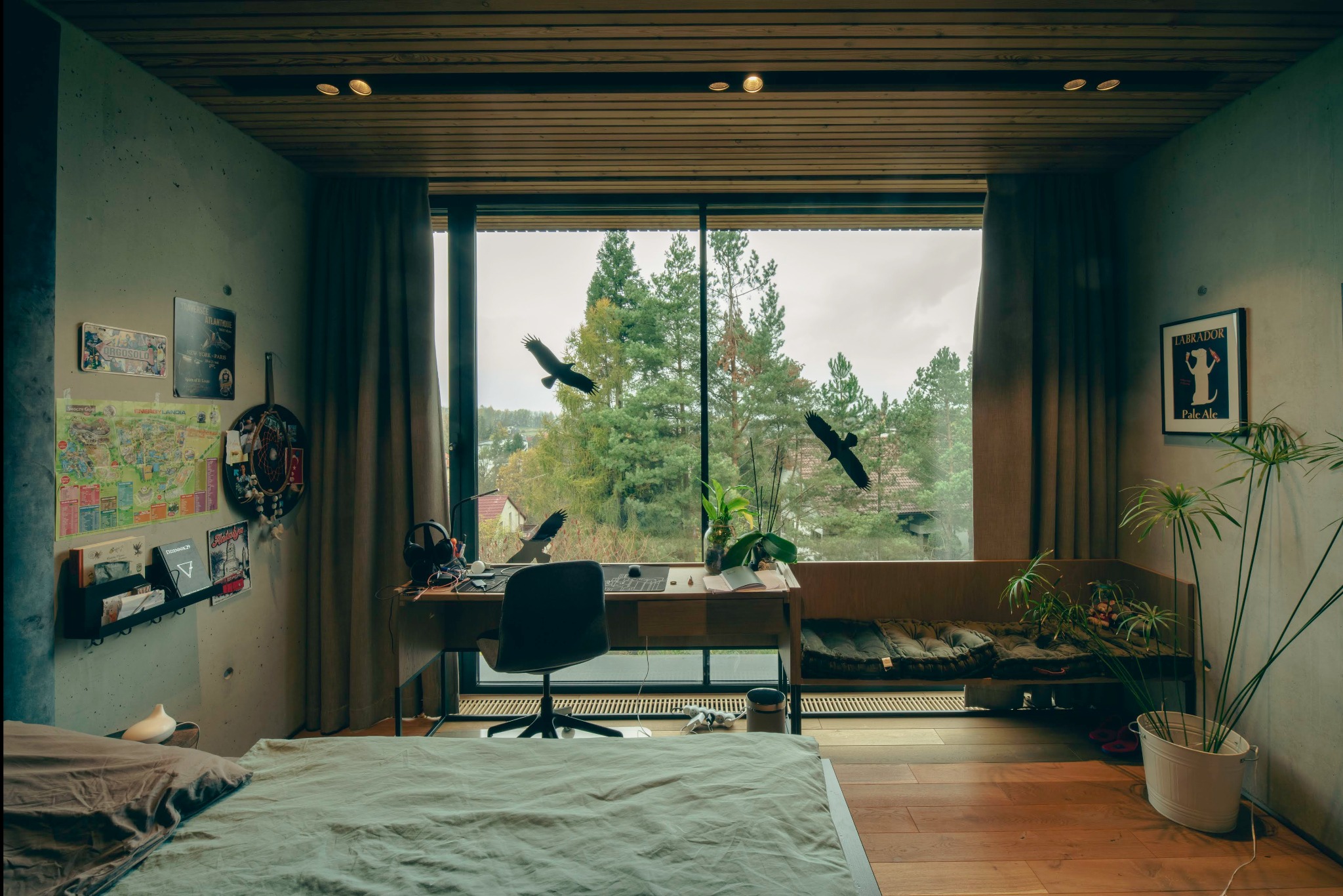 Pokój z miejscem do pracy przy dużym oknie z widokiem na drzewa, betonowe ściany ozdobione plakatami i mapą, drewniany sufit i podłoga, łóżko z zieloną pościelą na pierwszym planie.