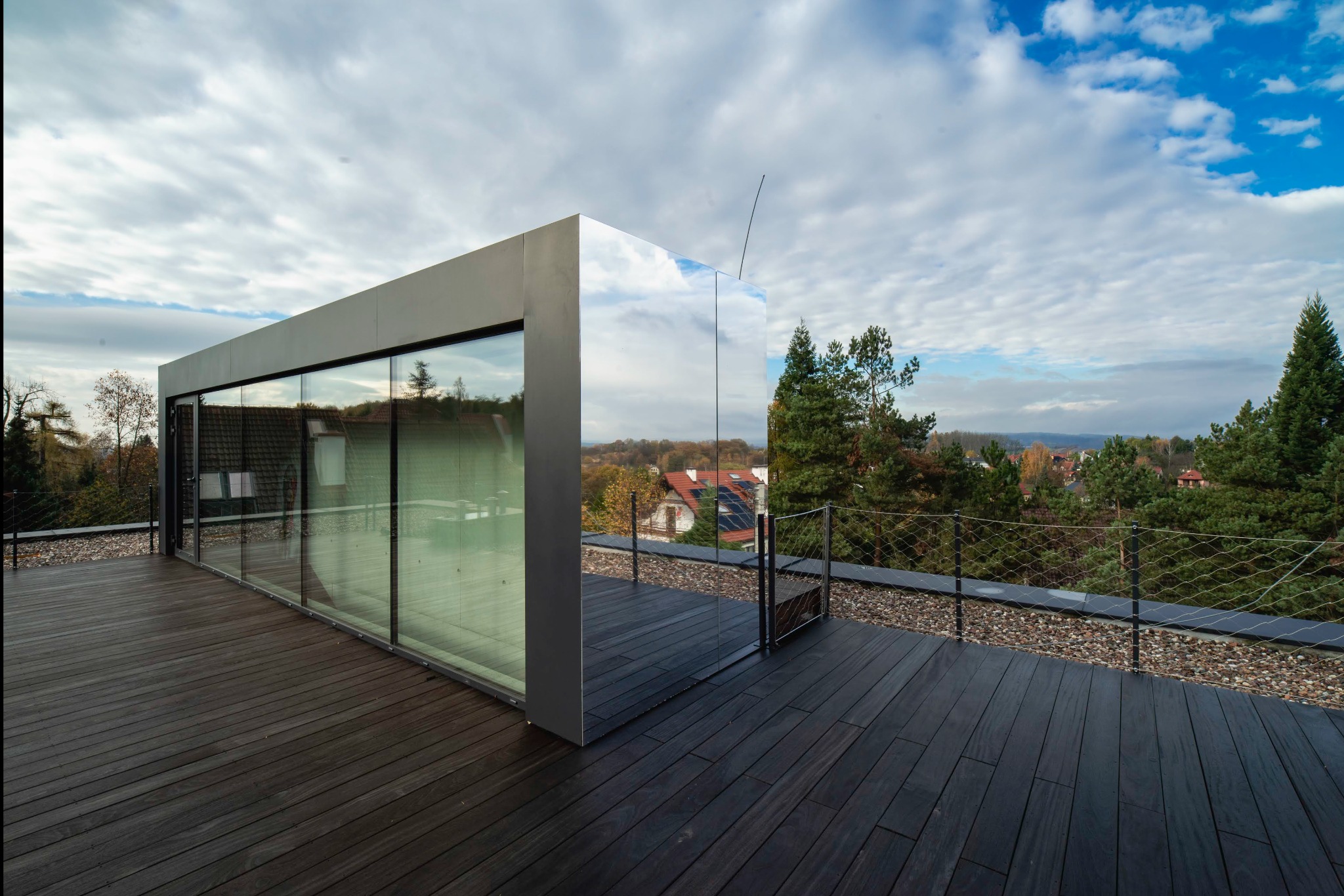 Nowoczesny taras na dachu z drewnianą podłogą i szklaną zabudową, odbijającą błękitne niebo i okoliczne drzewa.