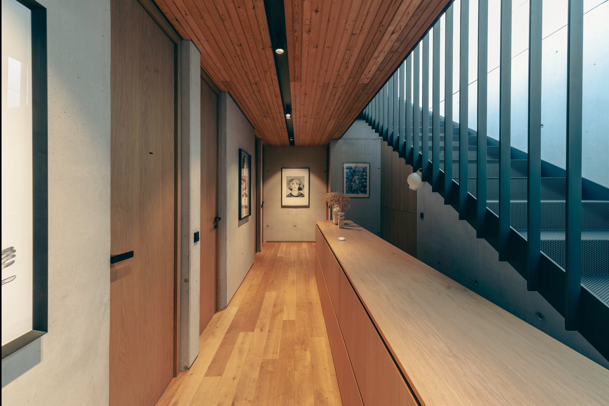 Wnętrze korytarza z drewnianą podłogą, boazerią sufitową i schodami z metalową balustradą, oświetlone punktowymi lampami sufitowymi, z minimalistycznymi drzwiami i dekoracjami ściennymi.