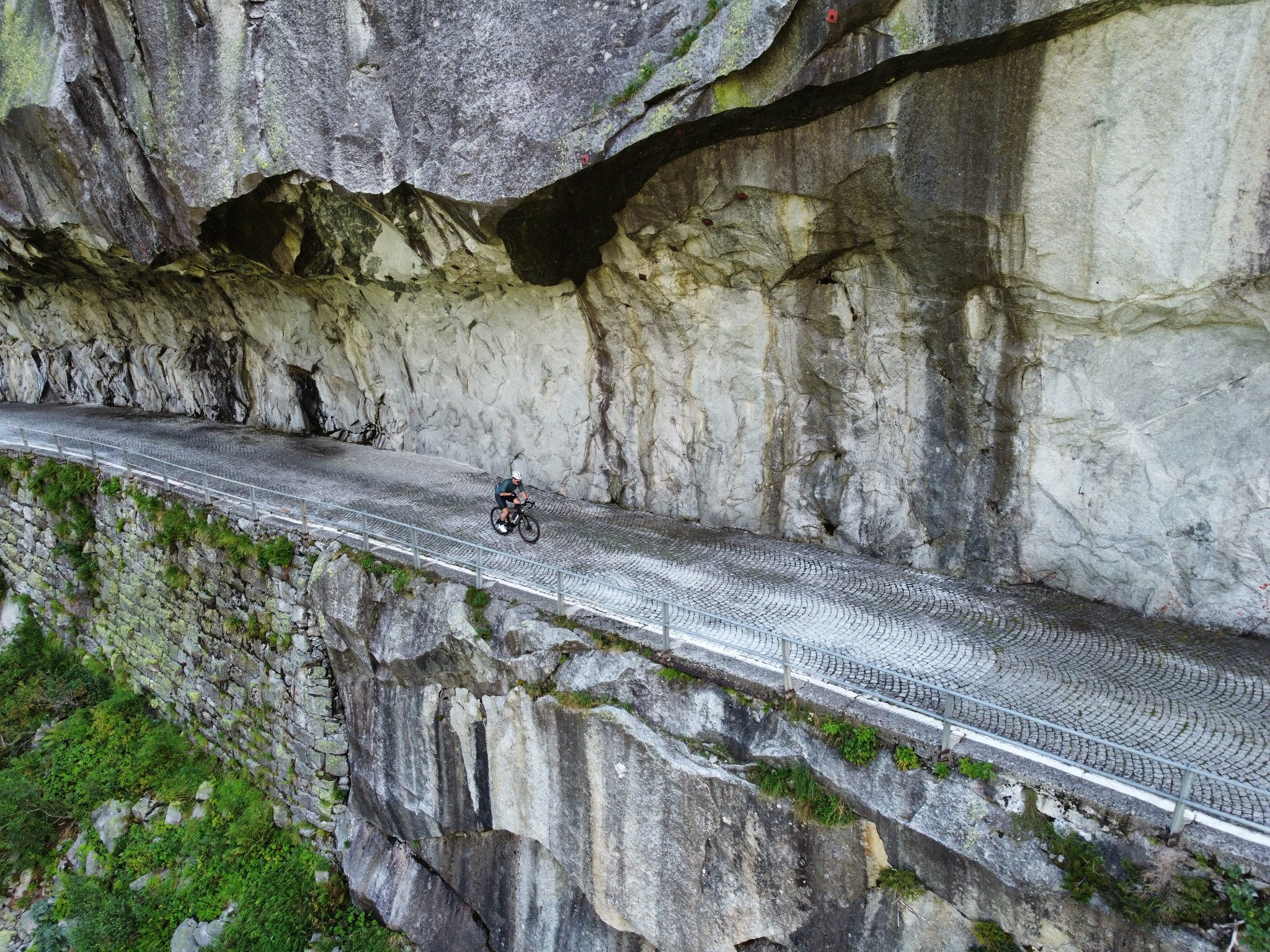 Kolarz na rowerze górskim jadący brukowaną drogą wykutą w skale, zabezpieczoną barierką, z widokiem z góry.
