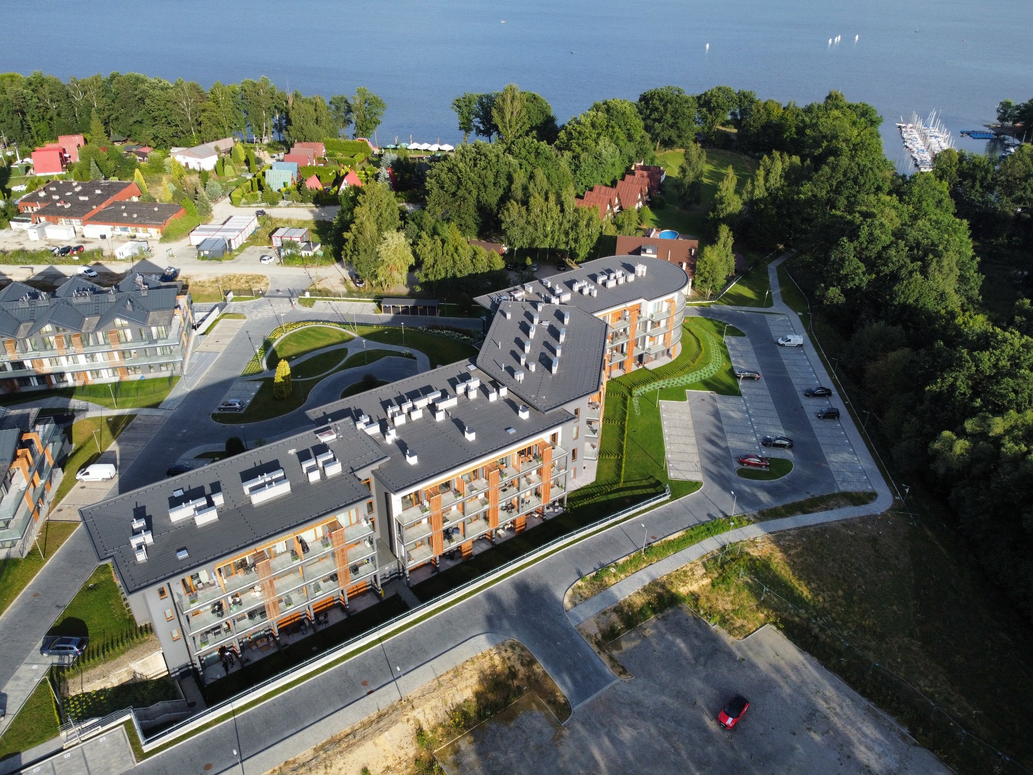 Panoramiczny widok z lotu ptaka na nowoczesny kompleks apartamentowy z balkonami, otoczony zielenią i jeziorem w tle.