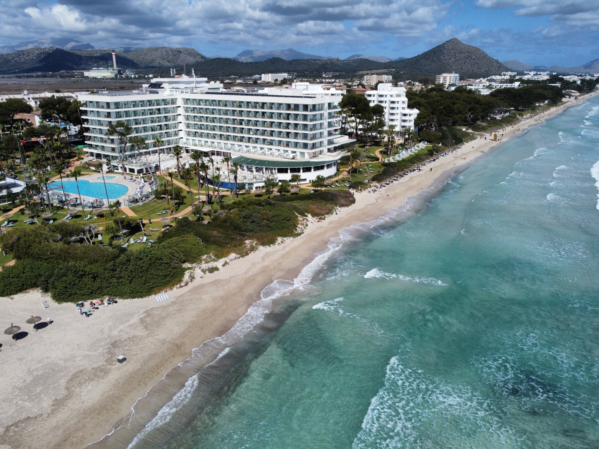 Panoramiczny widok z lotu ptaka na biały hotel z balkonami, basenem i palmami, położony przy piaszczystej plaży z turkusową wodą, w tle góry i zabudowania miejskie pod obłocznym niebem.