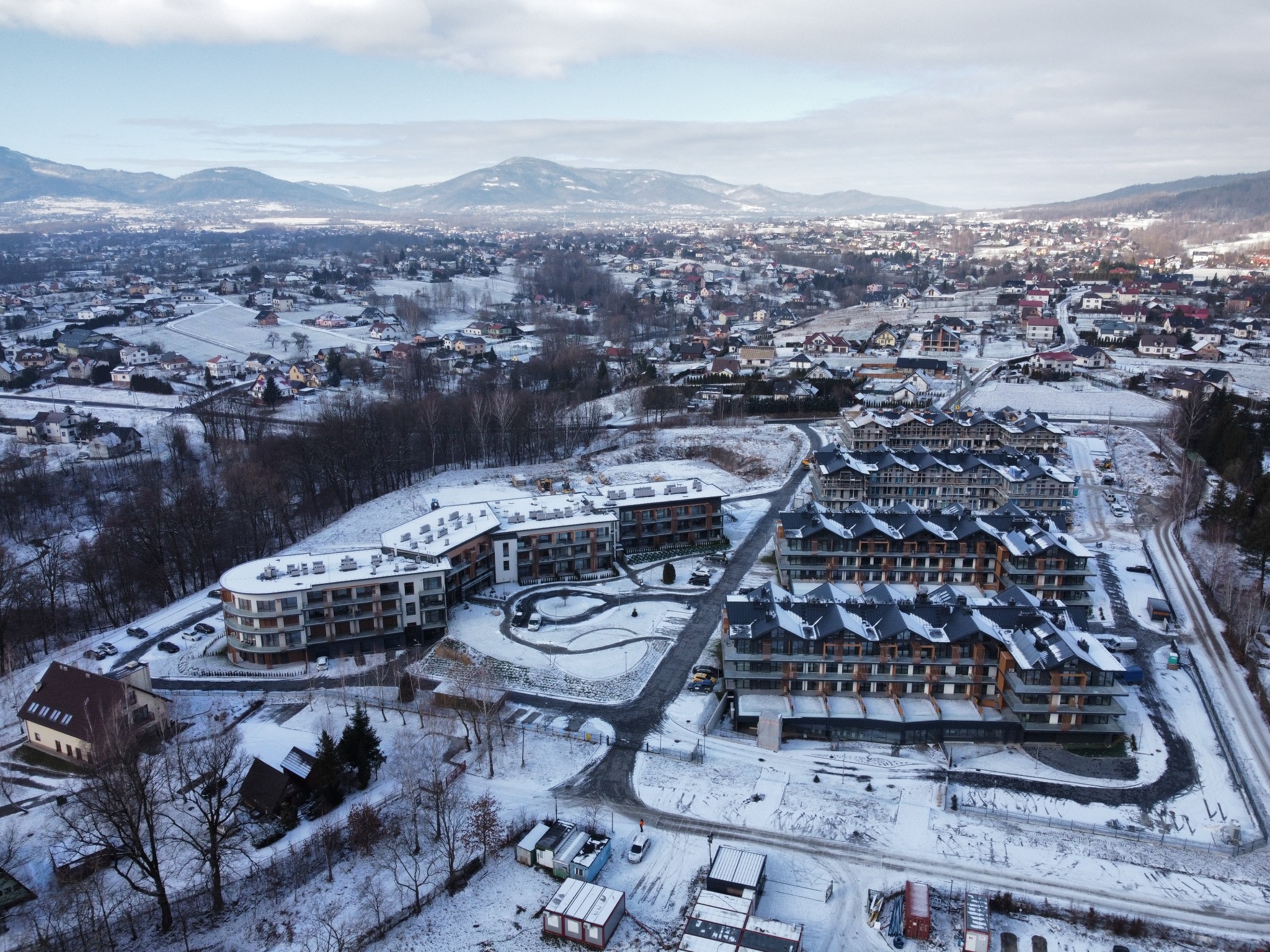 Zimowy krajobraz z lotu ptaka, przedstawiający kompleks apartamentowców z balkonami, pokrytych śniegiem, na tle gór i zabudowy miejskiej.