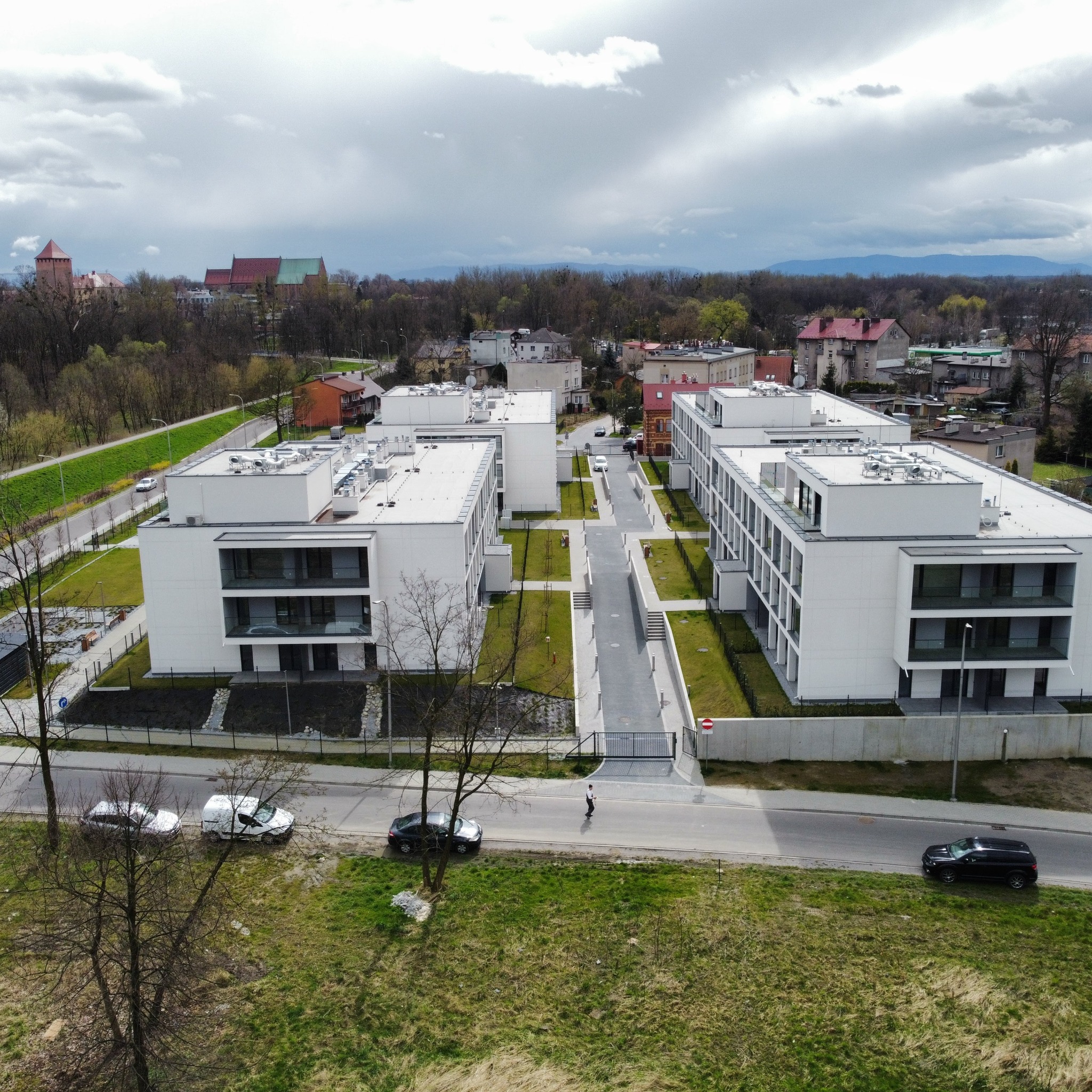 Widok z góry na kompleks nowoczesnych, białych budynków mieszkalnych z balkonami, otoczonych zielenią i drzewami, z widocznym w tle historycznym miastem pod pochmurnym niebem.