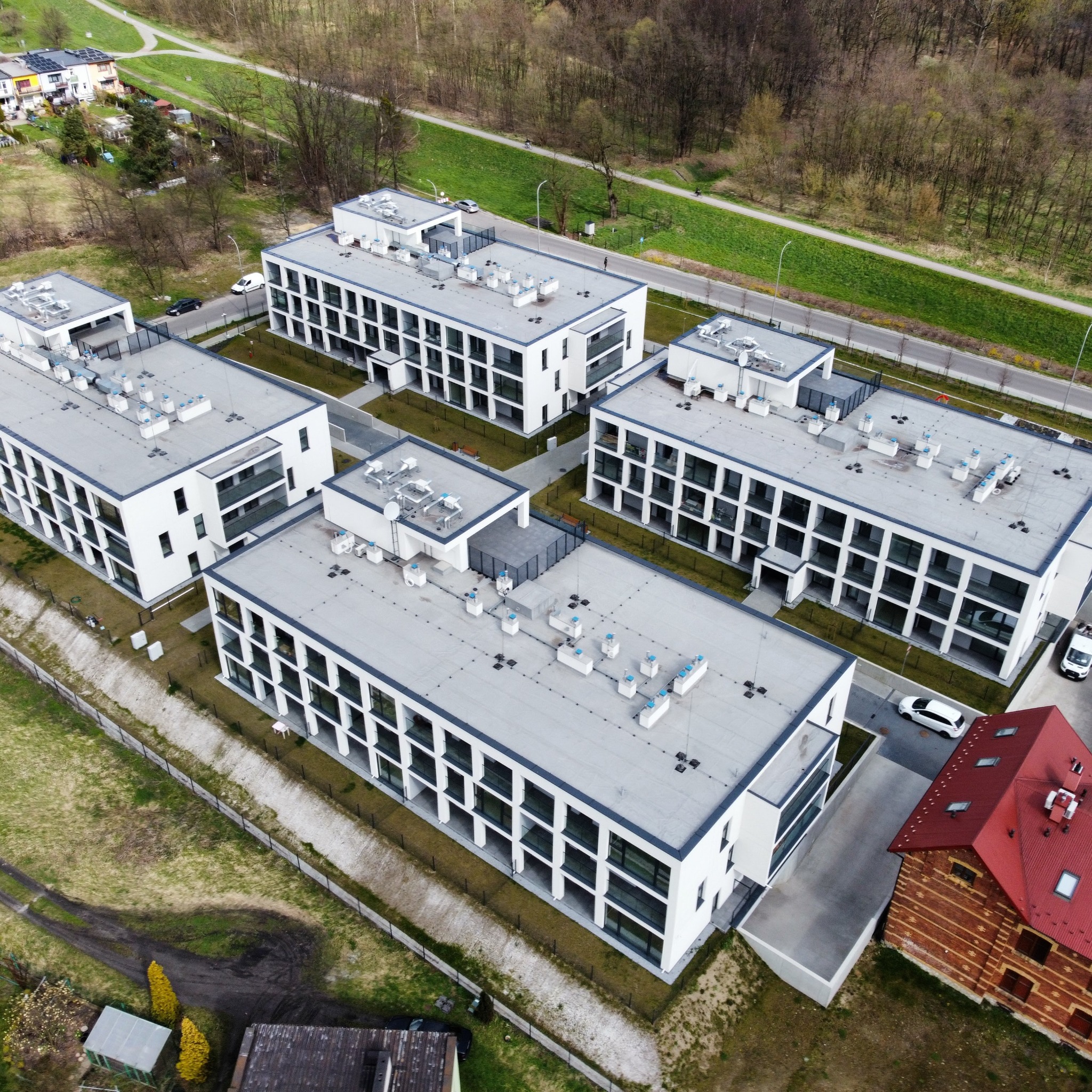Zdjęcie z drona przedstawia kompleks nowoczesnych, białych budynków apartamentowych z płaskimi dachami i licznymi balkonami, otoczonych zielenią, z widoczną starą ceglaną zabudową z czerwonym...