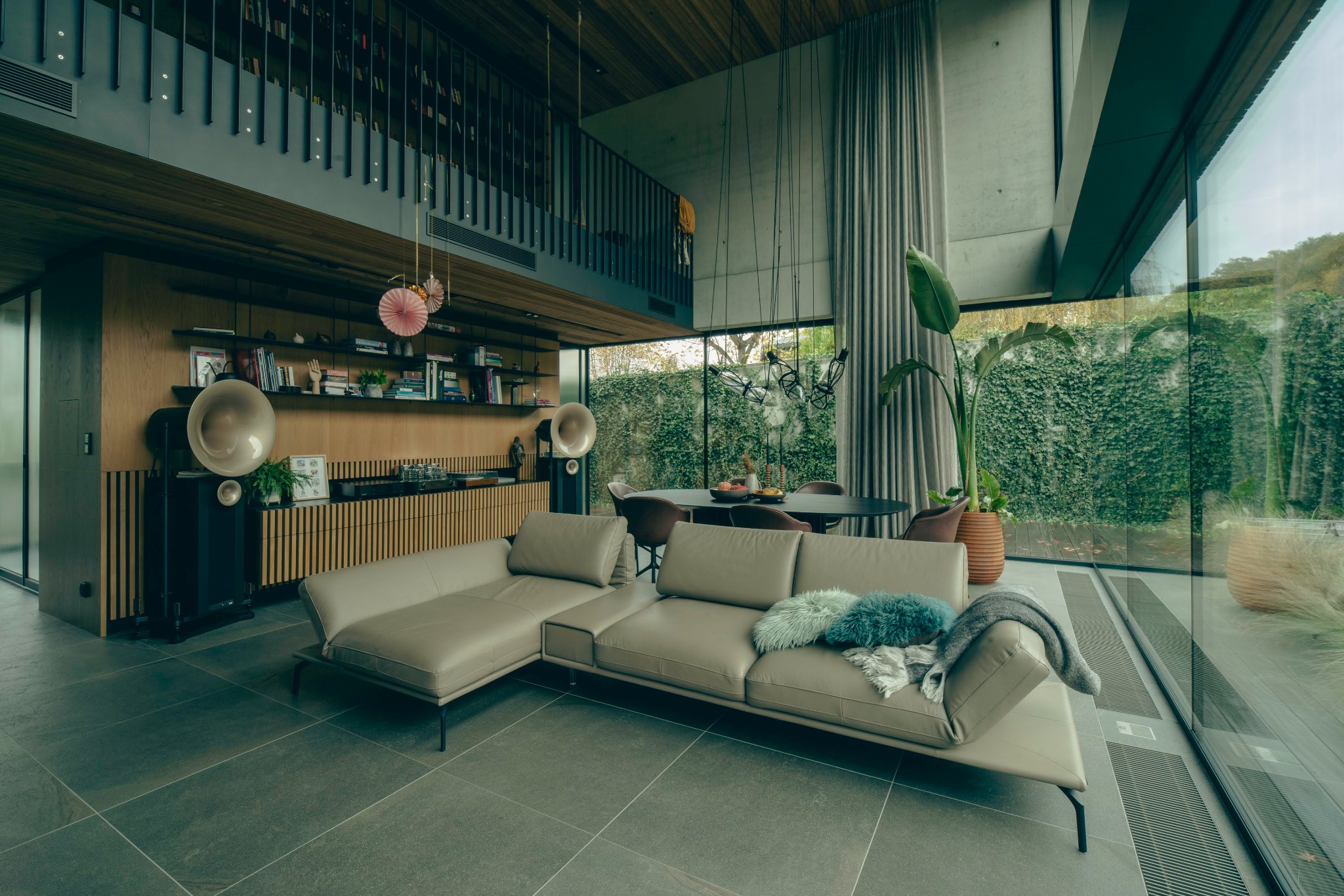 Nowoczesny salon z szarą podłogą, beżową sofą narożną, gramofonem tubowym i dużym oknem z widokiem na zielony ogród.