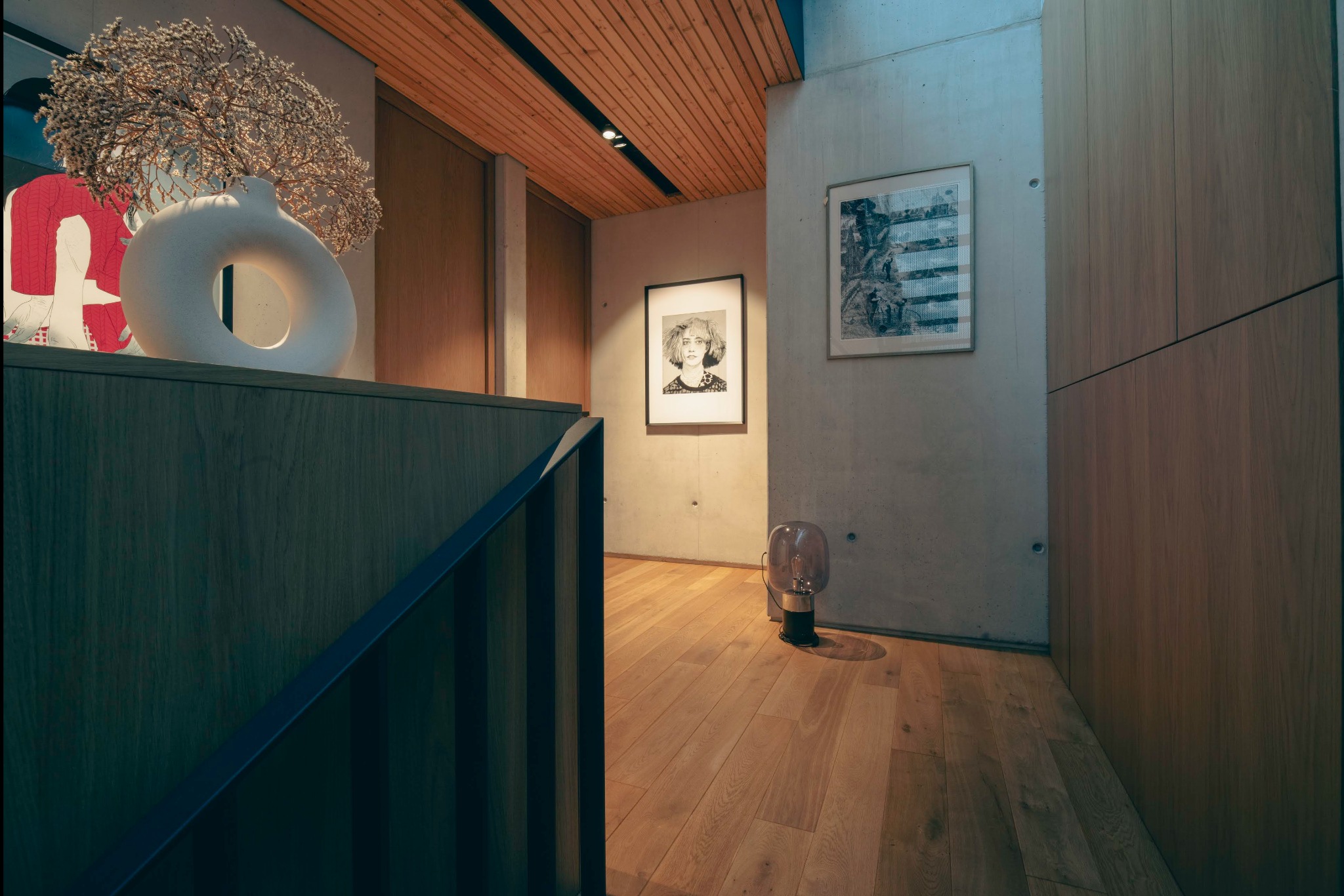Nowoczesny korytarz z drewnianą podłogą, betonową ścianą z grafikami i designerską lampą podłogową, uzupełniony dekoracyjnym wazonem z suszem na drewnianej balustradzie