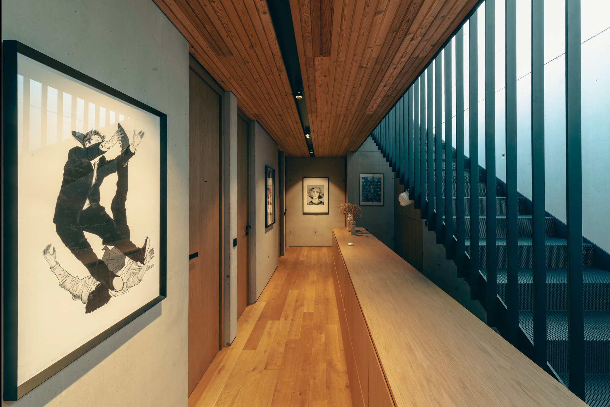 Wnętrze korytarza z drewnianą podłogą, sufitem i zabudową, betonowymi ścianami, obrazami w ramach i schodami z metalową balustradą.