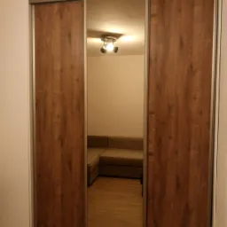 Trzydrzwiowa szafa przesuwna z lustrzanym panelem środkowym i okleiną imitującą drewno w ciepłym odcieniu brązu, odbijająca fragment salonu z narożną sofą.