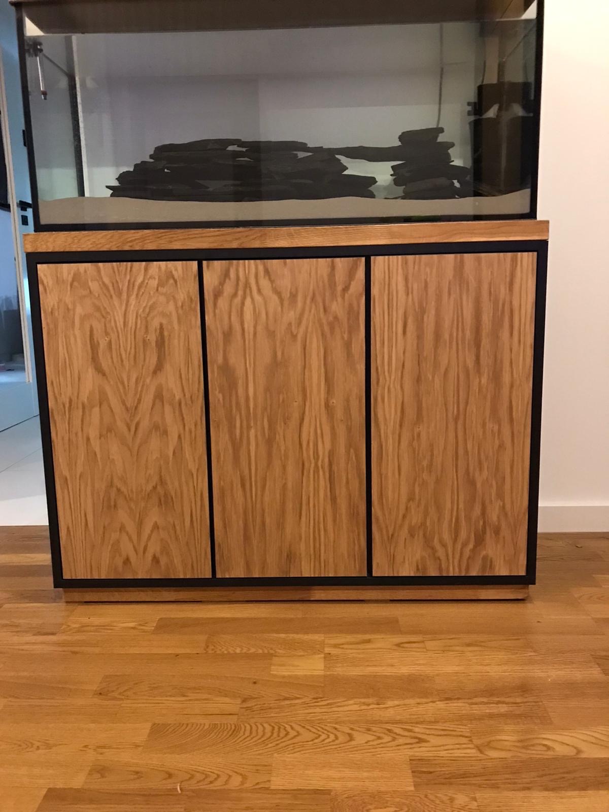 Akrylowe akwarium z czarnymi skałami i beżowym podłożem, umieszczone na szafce z trzema drewnianymi drzwiczkami z czarną ramą, stojącej na parkiecie.