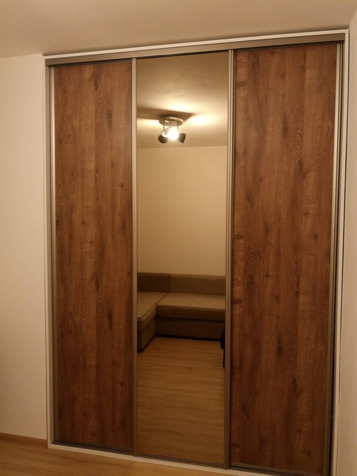 Trzydrzwiowa szafa przesuwna z lustrzanym panelem środkowym i okleiną imitującą drewno w ciepłym odcieniu brązu, odbijająca fragment salonu z narożną sofą.