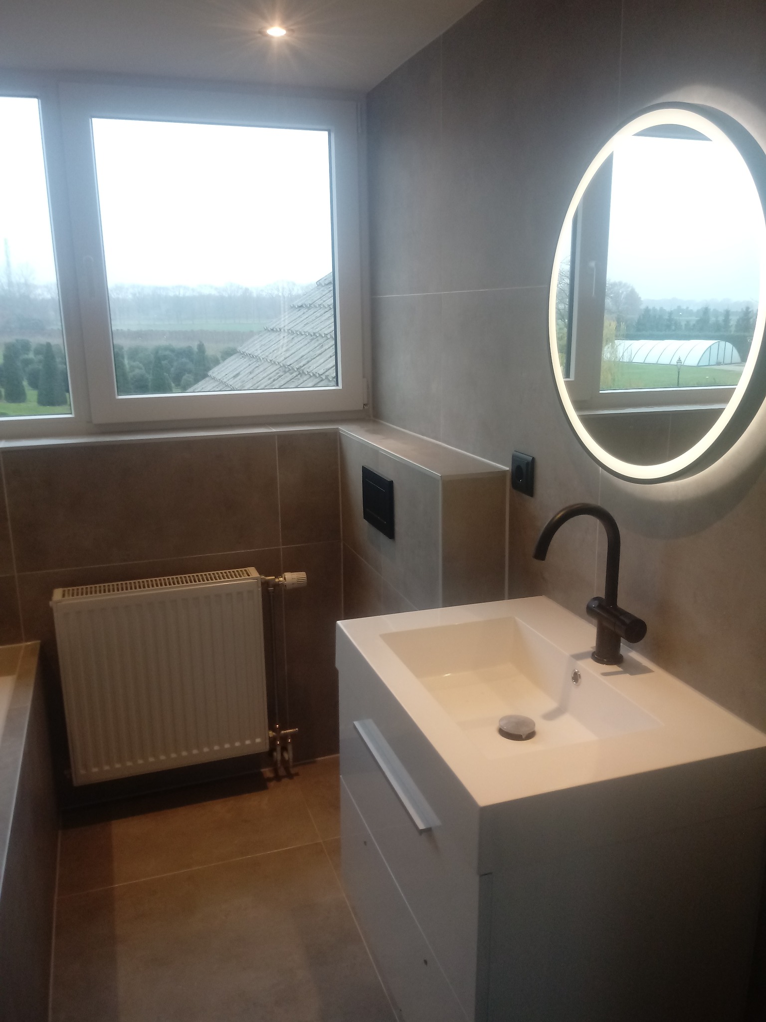 Nowoczesna łazienka w szaro-brązowej tonacji z podświetlanym lustrem, białą umywalką z szafką i grzejnikiem pod oknem z widokiem na zewnątrz.