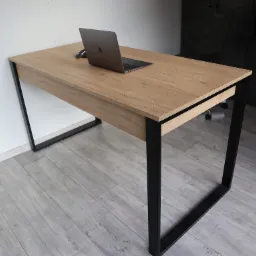 Biurko z blatem imitującym jasne drewno i czarnymi metalowymi nogami, na blacie laptop i myszka komputerowa, w tle fragment szafy.