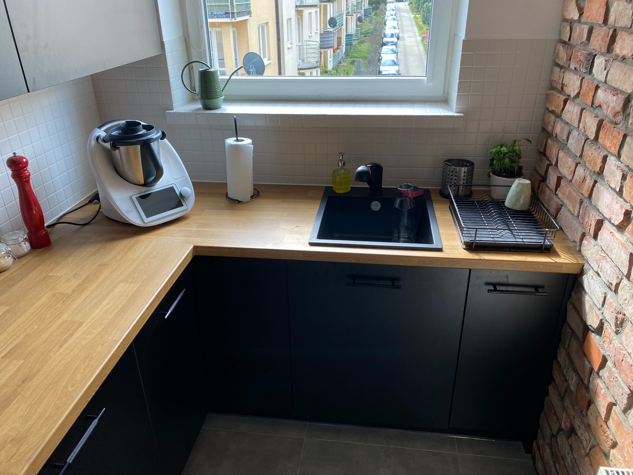 Fragment kuchni z drewnianym blatem, czarnym zlewem i szafkami, na blacie robot kuchenny, widok z okna na ulicę, ściana z cegły.