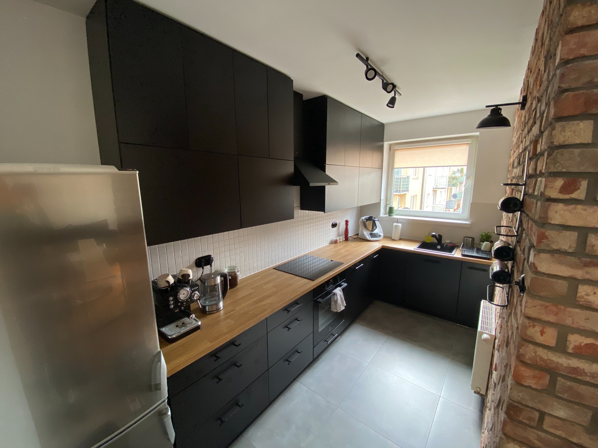 Nowoczesna kuchnia z czarnymi szafkami, drewnianym blatem, białymi płytkami na ścianie i ceglaną ścianą boczną z wieszakiem na wino.