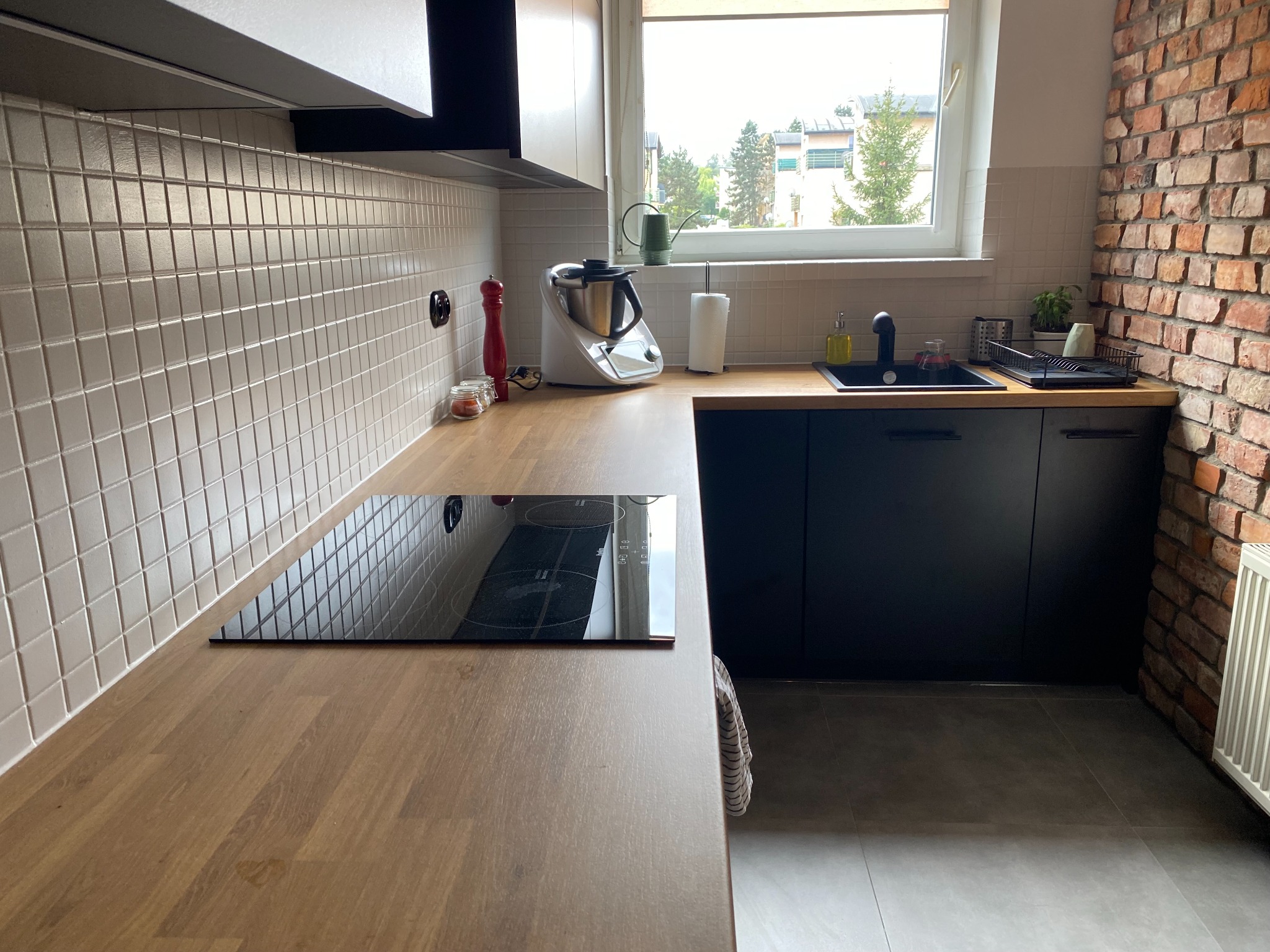 Nowoczesna kuchnia z drewnianym blatem, czarnymi szafkami i płytą indukcyjną, uzupełniona o ceglaną ścianę i widok za oknem.