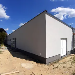 Elewacja budynku po remoncie, ściany w jasnym odcieniu szarości, widoczne białe drzwi i okna, teren wokół budynku częściowo zagospodarowany.