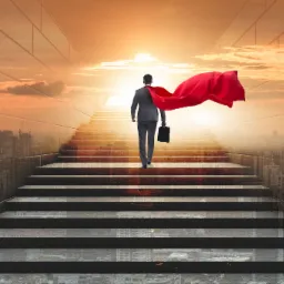 Mężczyzna w garniturze i czerwonej pelerynie wchodzi po schodach w kierunku wschodzącego słońca, niosąc aktówkę.