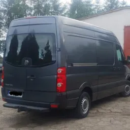 Szary Volkswagen Crafter z widocznym hakiem holowniczym, zaparkowany na zewnątrz przy budynku magazynowym.
