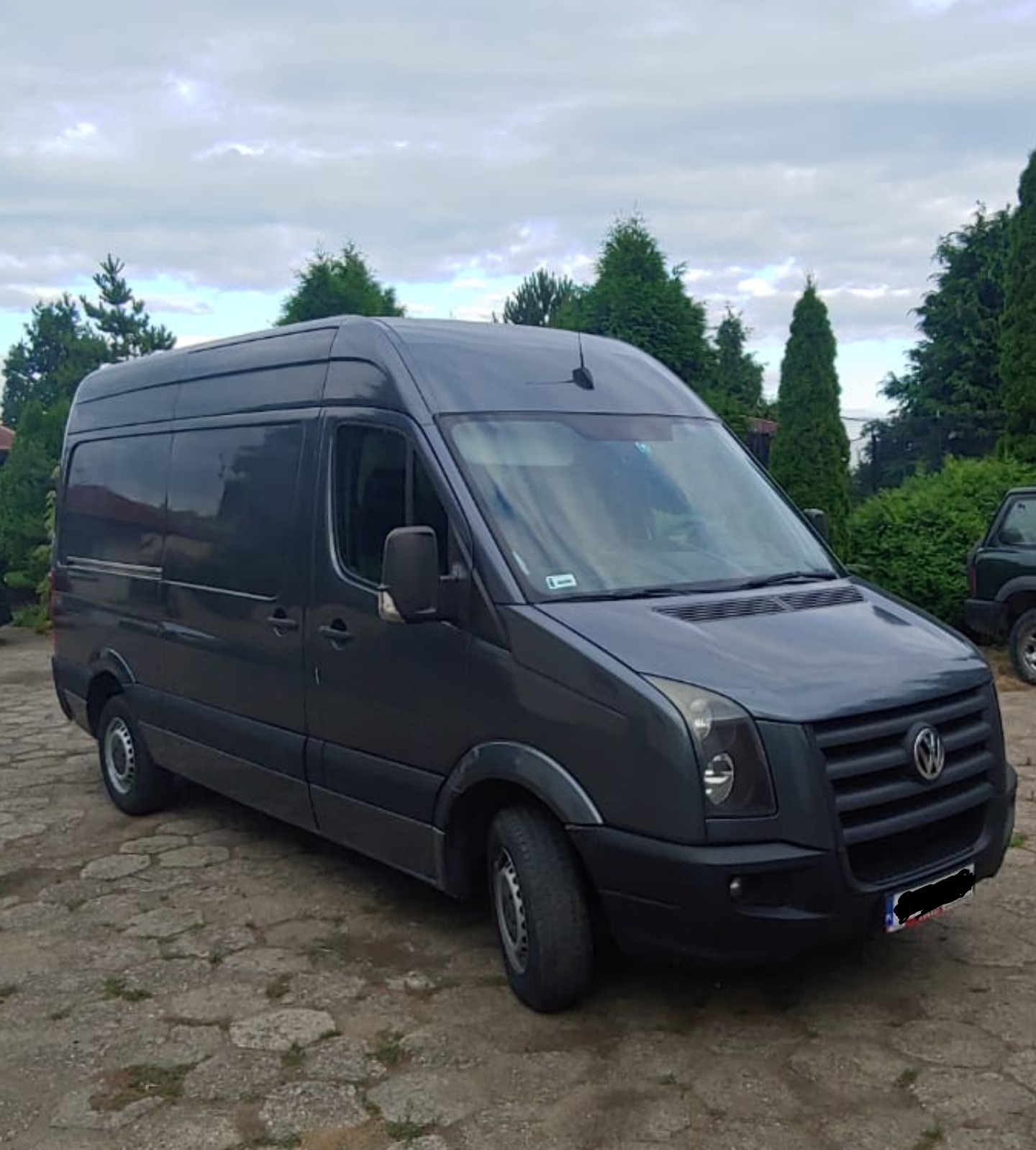 Szary, wysoki bus Volkswagen Crafter na brukowanym podjeździe, z tłem zielonych drzew i pochmurnego nieba.