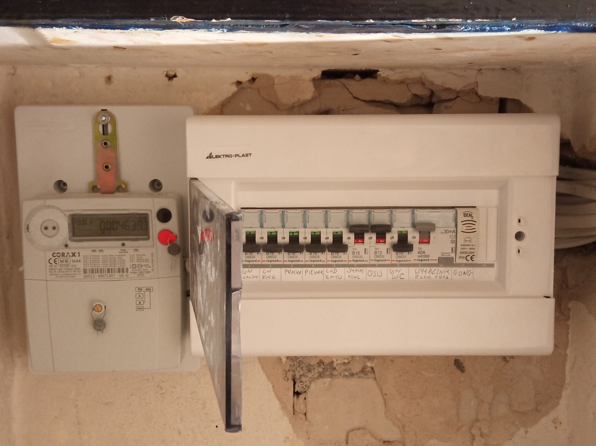 Zamontowana skrzynka elektryczna Elektro-Plast z otwartymi drzwiczkami obok licznika CORAX1 w surowym wnętrzu ze śladami tynku i przewodami.