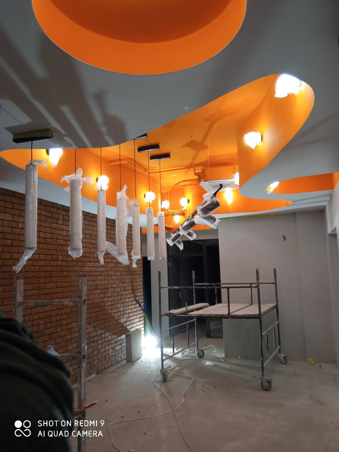 Montaż oświetlenia: wiszące lampy w folii ochronnej pod pomarańczowym sufitem, widoczny fragment ceglanej ściany i rusztowanie.