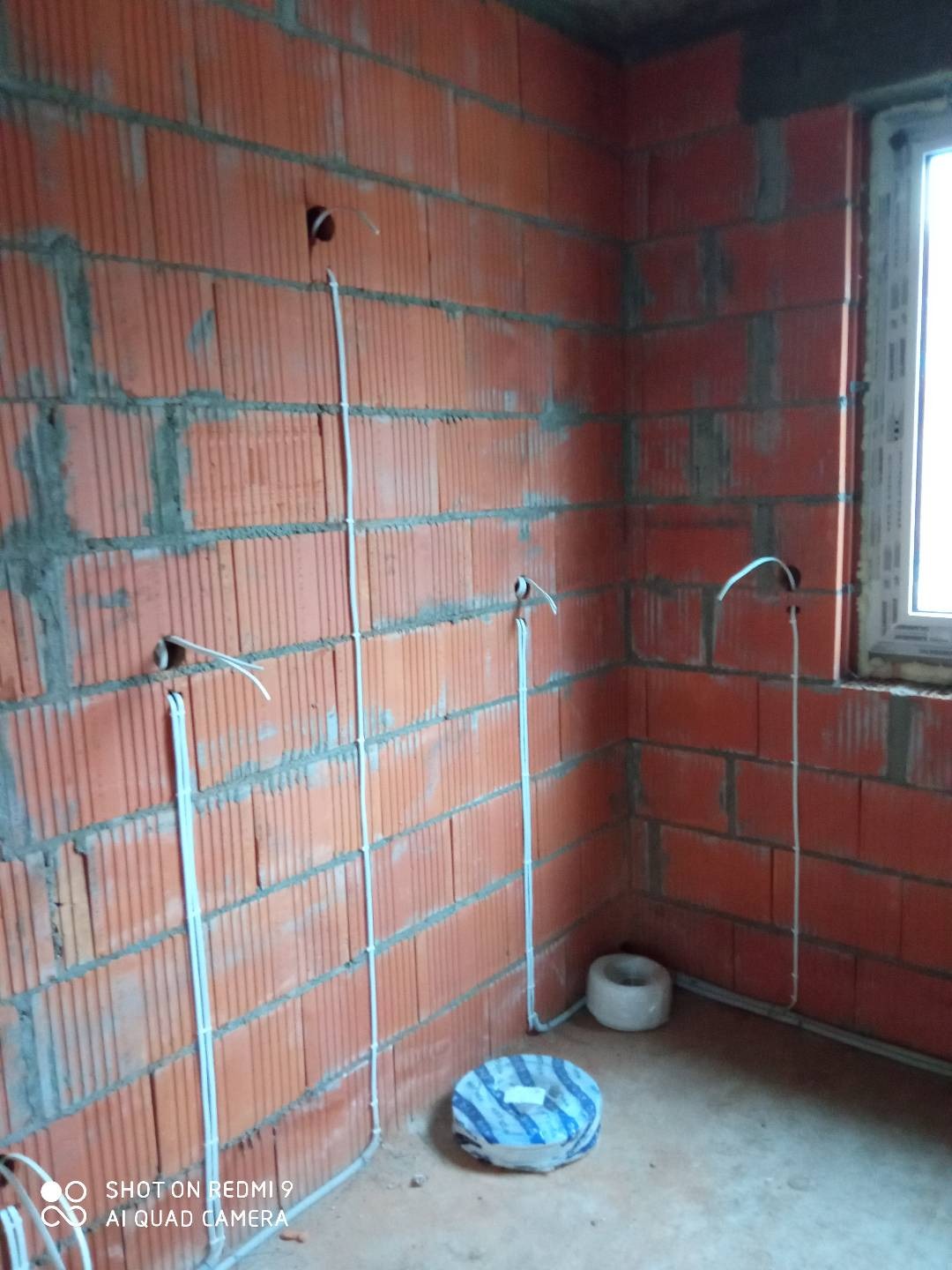 Świeżo murowana ściana z pustaków ceramicznych z poprowadzonymi przewodami elektrycznymi w białych rurkach instalacyjnych, widoczne puszki instalacyjne i zwój kabla na podłodze.