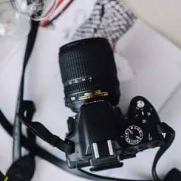 Czarny aparat Nikon z obiektywem 18-105mm leży na otwartym magazynie modowym obok żarówki. Ujęcie z góry, delikatne światło.