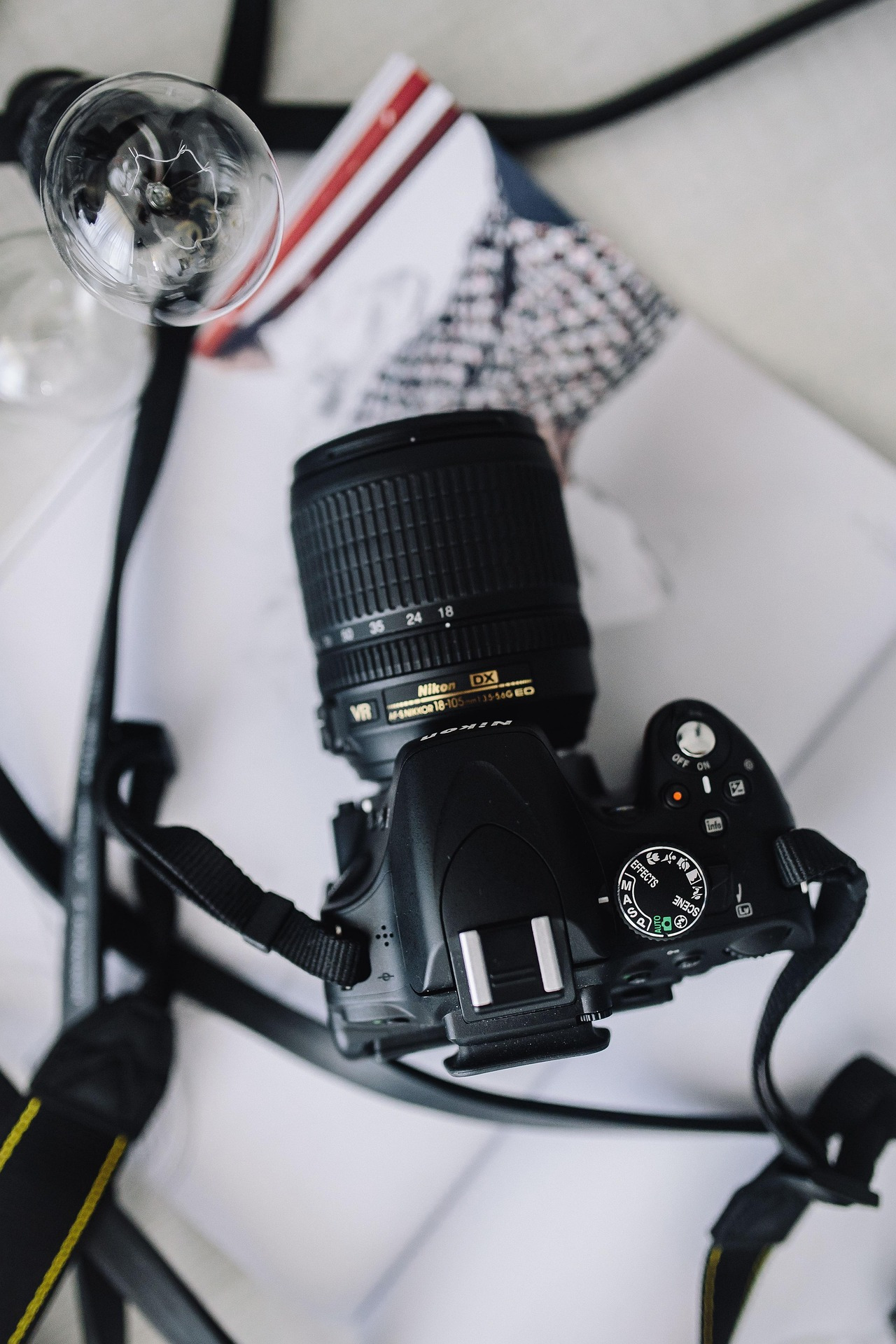Czarny aparat Nikon z obiektywem 18-105mm leży na otwartym magazynie modowym obok żarówki. Ujęcie z góry, delikatne światło.