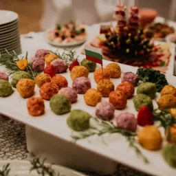 Kolorowe kulki serowe (zielone, pomarańczowe, fioletowe) na białym prostokątnym talerzu, udekorowane gałązkami rozmarynu i truskawkami, flaga wbita w jedną z kulek, w tle inne dania i stos talerzy,...