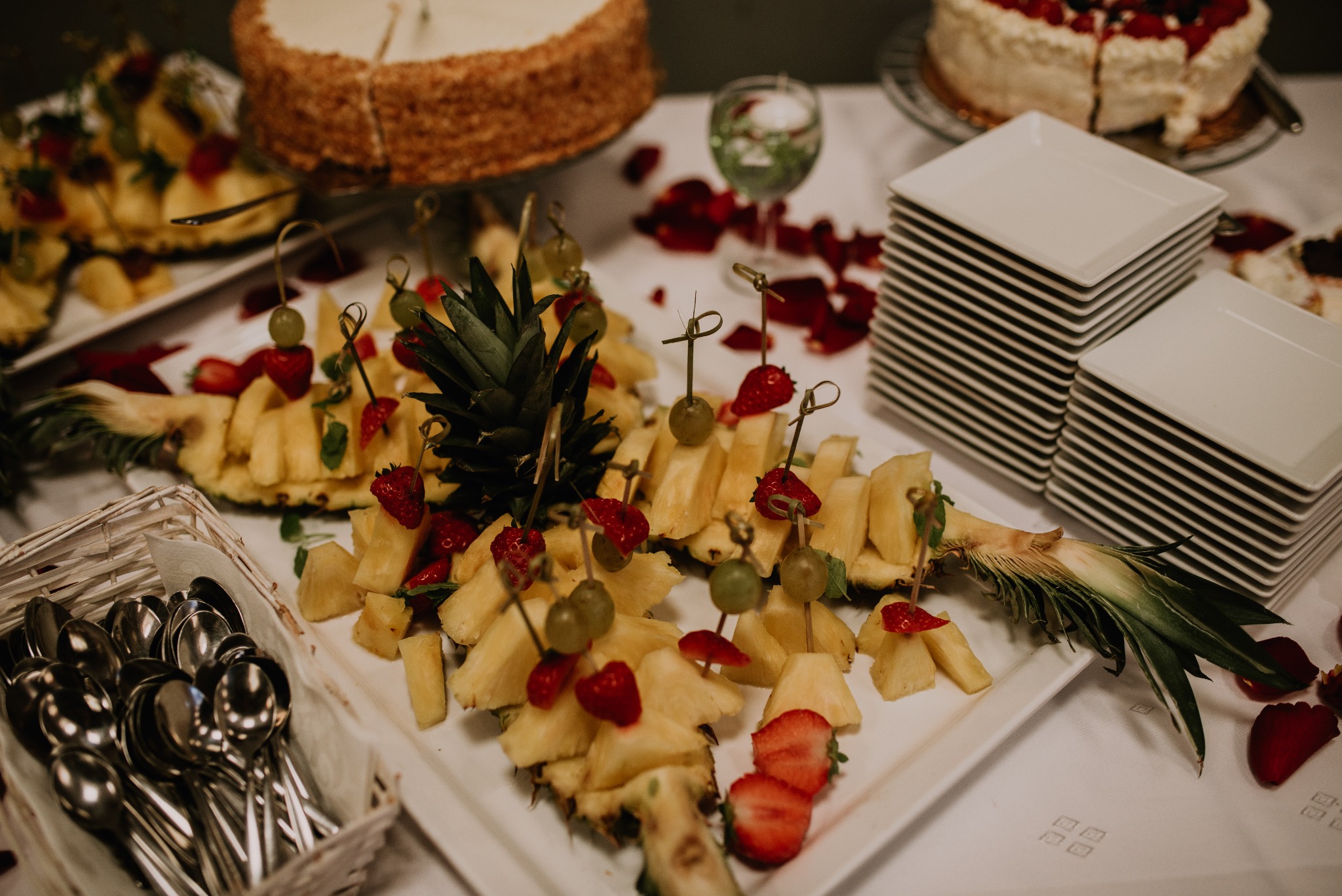 Udekorowany stół z przekąskami: ananas pokrojony w kostkę z owocami na wykałaczkach, torty w tle, stosy białych talerzy i koszyk ze sztućcami.
