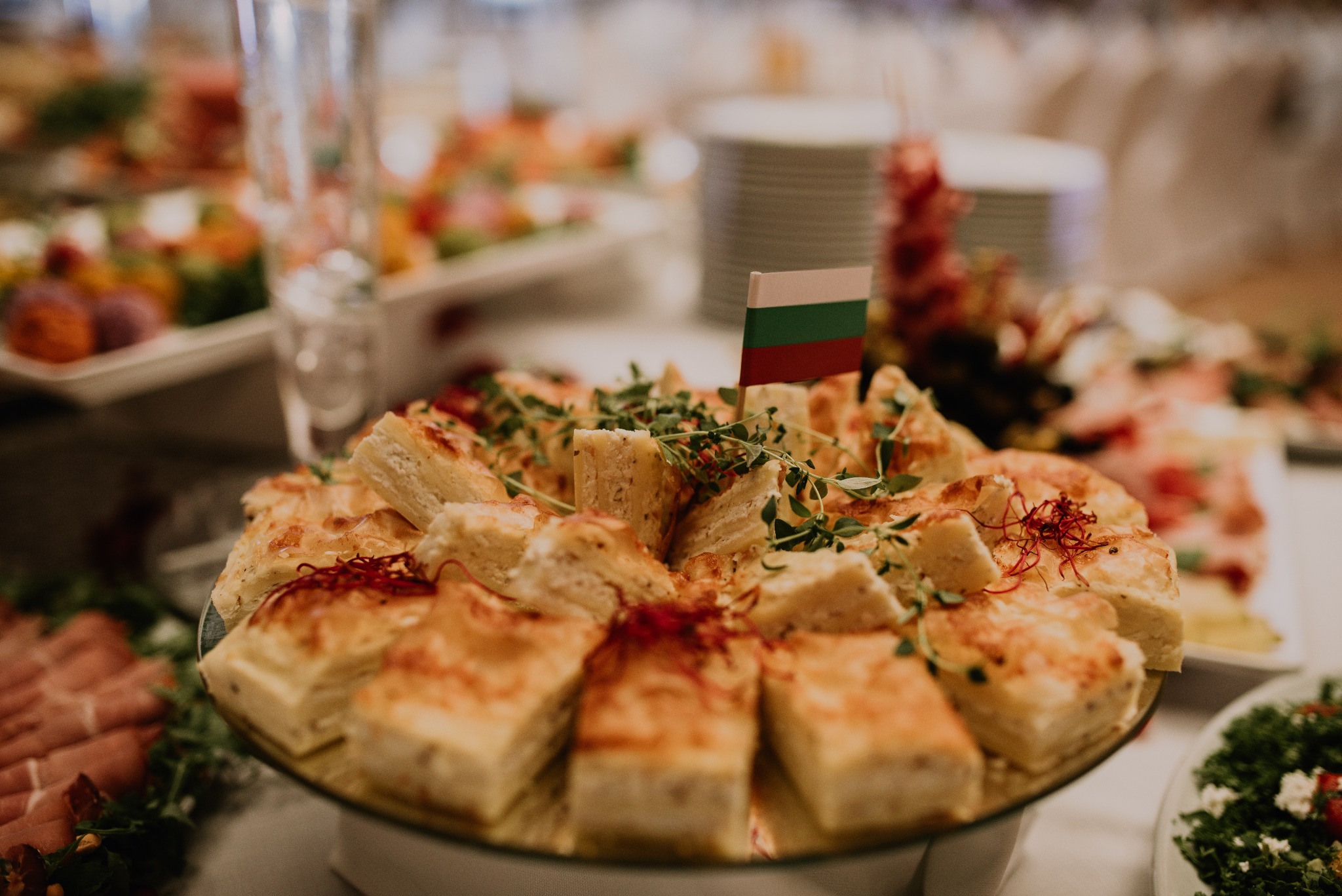 Elegancko podane kawałki ciasta na szklanym talerzu, udekorowane ziołami i małą flagą Bułgarii, w tle inne przekąski i napoje na stole.