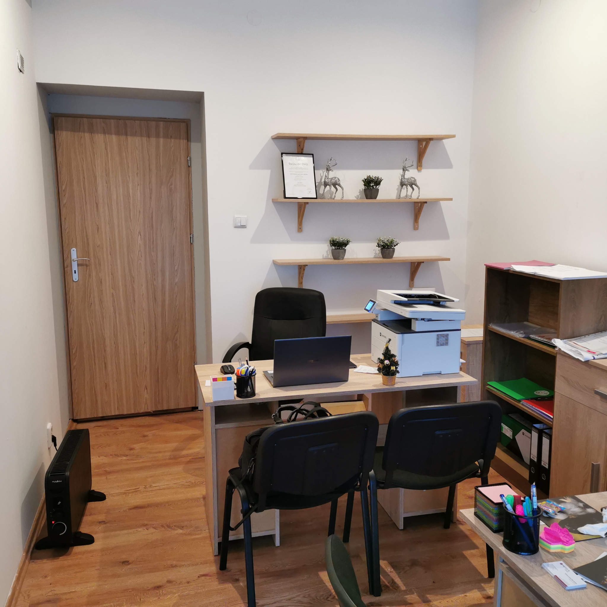 Biuro z biurkiem, laptopem, drukarką, półkami z dekoracjami i grzejnikiem elektrycznym przy ścianie.