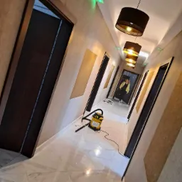 Korytarz po remoncie z marmurową podłogą, wzorzystymi ścianami i wiszącymi lampami, widoczny odkurzacz i osoba w oddali.