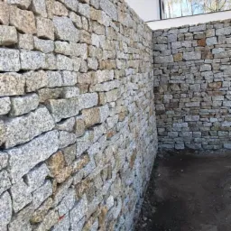 Dwie ściany z nieregularnej kostki granitowej w odcieniach szarości i beżu, ułożone pod kątem prostym, z widocznym fragmentem budynku w tle.