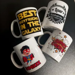 Cztery białe kubki z różnymi nadrukami leżące na ciemnym tle: napis 'Best Boyfriend in the Galaxy', herb z napisem 'Laur Konkubenta', postać superbohatera z imieniem 'Domixxx', oraz komiksowe...