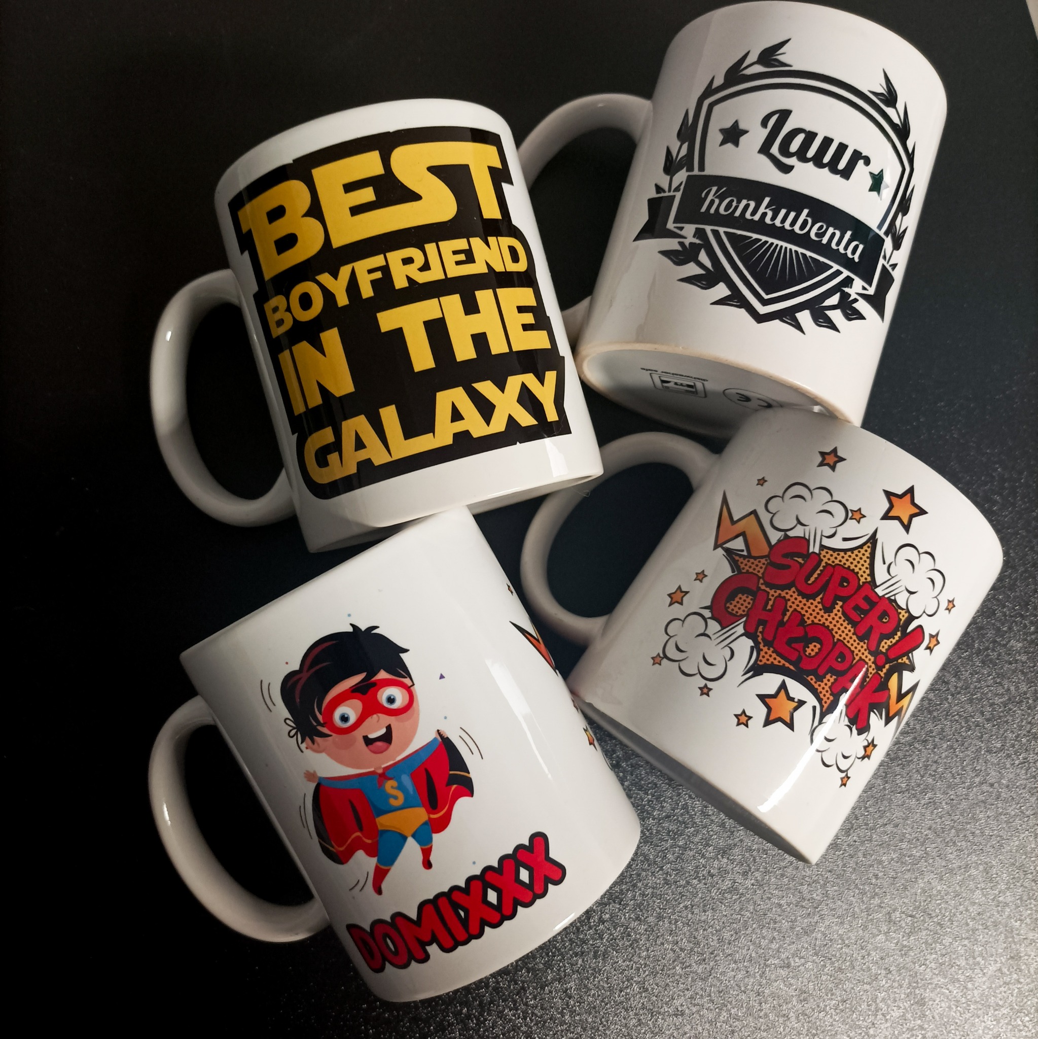 Cztery białe kubki z różnymi nadrukami leżące na ciemnym tle: napis 'Best Boyfriend in the Galaxy', herb z napisem 'Laur Konkubenta', postać superbohatera z imieniem 'Domixxx', oraz komiksowe...