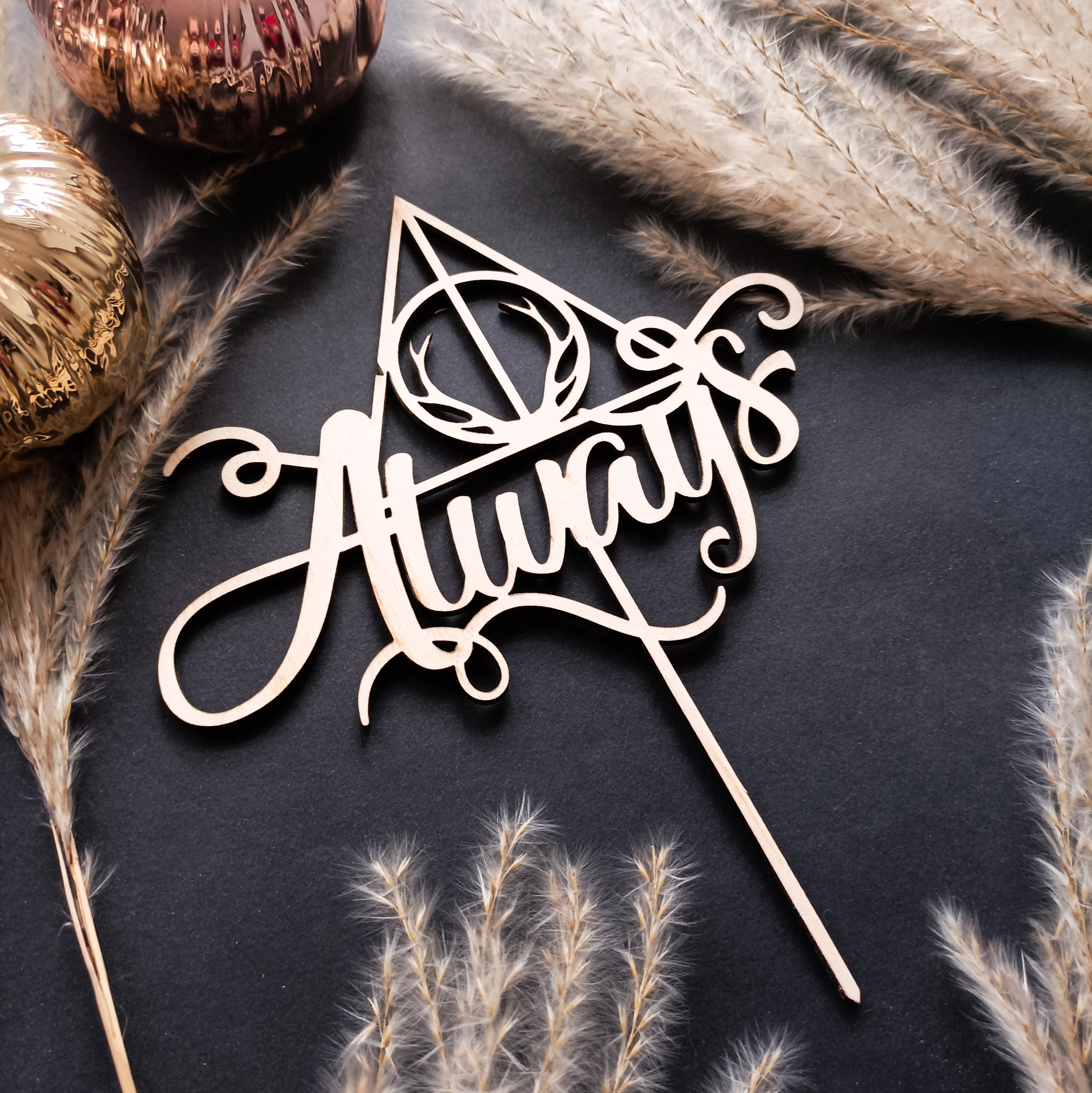 Drewniany topper na tort z napisem 'Always' i symbolem Insygniów Śmierci, otoczony suszonymi trawami i złotymi bombkami na ciemnym tle.