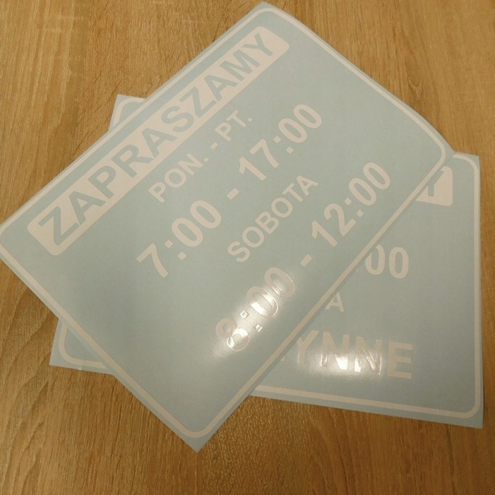 Dwie jasnoniebieskie naklejki z białym napisem informującym o godzinach otwarcia: 'Zapraszamy Pon.-Pt. 7:00-17:00 Sobota 8:00-12:00' leżące na drewnianym blacie.