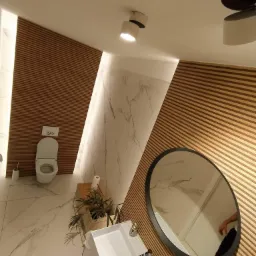 Nowoczesna łazienka z drewnianą boazerią, marmurową ścianą, owalnym lustrem i białą umywalką, oświetlona punktowymi lampami sufitowymi.