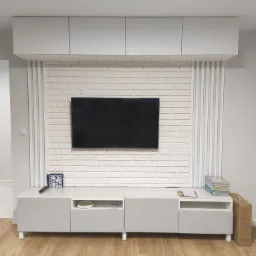 Nowoczesny salon z białą cegłą dekoracyjną na ścianie za telewizorem, szare szafki wiszące i pod telewizorem, drewniana podłoga.