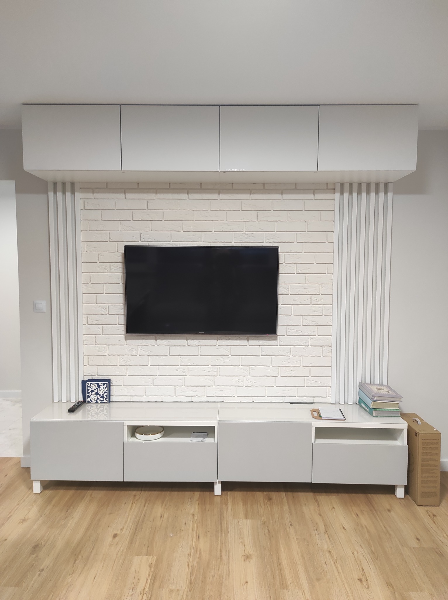 Nowoczesny salon z białą cegłą dekoracyjną na ścianie za telewizorem, szare szafki wiszące i pod telewizorem, drewniana podłoga.