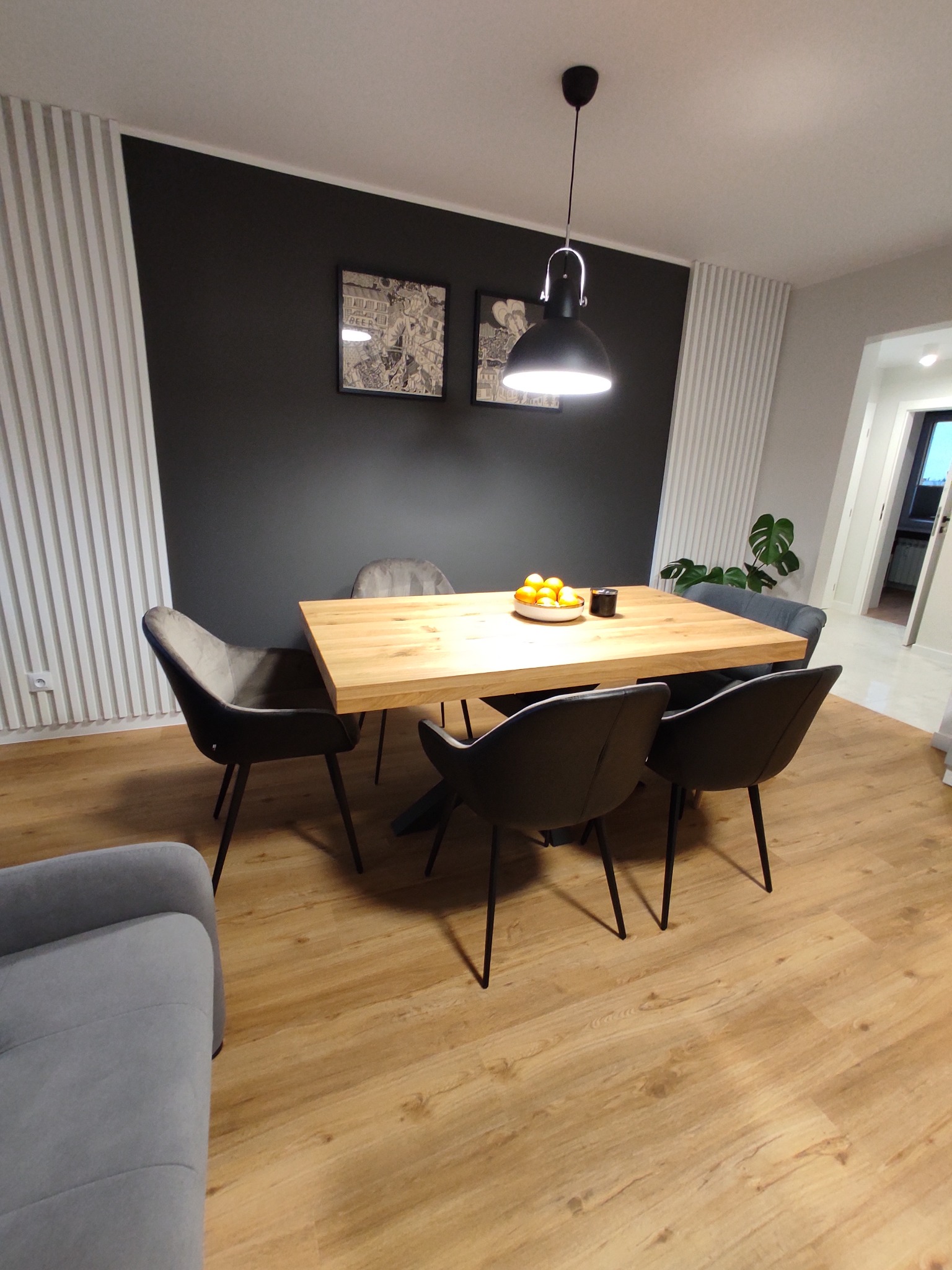 Jadalnia w nowoczesnym stylu: drewniany stół z owocami, czarne krzesła na tle ciemnej ściany z dekoracyjnymi panelami, oświetlona wiszącą lampą.