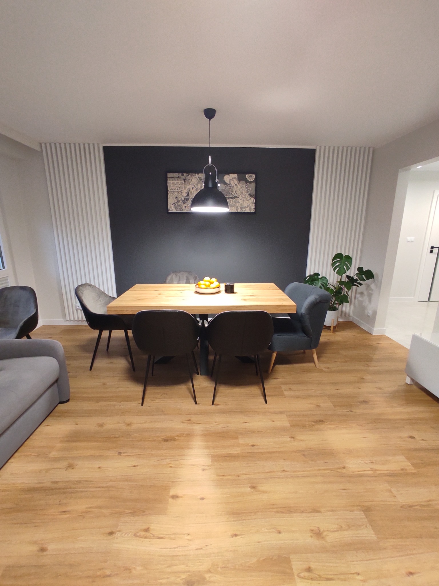 Nowoczesna jadalnia z drewnianym stołem, czarnymi krzesłami i lampą wiszącą, ściana akcentowa w kolorze grafitowym z białymi panelami dekoracyjnymi po bokach, podłoga z paneli imitujących drewno.