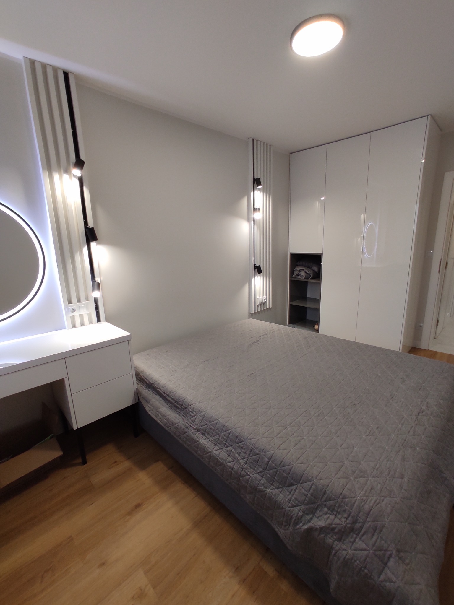 Nowoczesna sypialnia z szarą pościelą na łóżku, białymi szafami w zabudowie z półkami i toaletką z okrągłym lustrem podświetlanym LED, oświetlenie punktowe na ścianie.